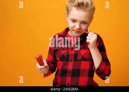 Junge isst trockene Würstchen. Schöner lächelnder kaukasischer Junge im roten Hemd auf dem gelben Hintergrund. Horizontal, gerade. Stockfoto