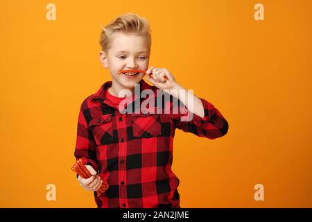 Junge isst trockene Würstchen. Schöner lächelnder kaukasischer Junge im roten Hemd auf dem gelben Hintergrund. Horizontal, gerade. Stockfoto