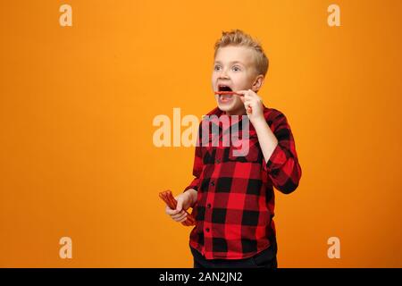 Junge isst trockene Würstchen. Schöner lächelnder kaukasischer Junge im roten Hemd auf dem gelben Hintergrund. Horizontal, gerade. Stockfoto