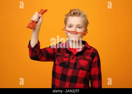 Junge isst trockene Würstchen. Schöner lächelnder kaukasischer Junge im roten Hemd auf dem gelben Hintergrund. Horizontal, gerade. Stockfoto