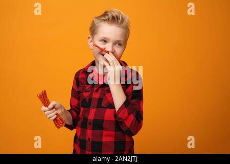 Junge isst trockene Würstchen. Schöner lächelnder kaukasischer Junge im roten Hemd auf dem gelben Hintergrund. Horizontal, gerade. Stockfoto