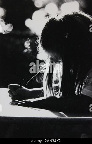 Feine Siebziger Schwarz-Weiß-Extremfotografie von Mädchen, die an einem Schreibtisch mit Hintergrundbeleuchtung schreiben Stockfoto