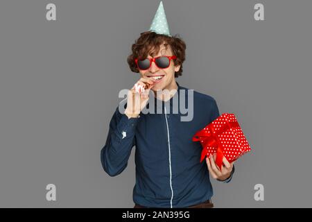 Feier Konzept. Teenager Junge in Geburtstag Kappe und Sonnenbrille isoliert auf Grau mit Geschenk blasen Party horn Lachen verspielt Stockfoto