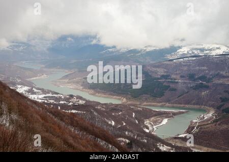 Sanfter, nebliger Blick auf die Flecken des Sonnenlichts, die auf schneebedeckten Kiefernwäldern und in der bedeckten Landschaft rund um den schönen, mäandrierenden, zugefrorenen See Zavoj in Ser scheinen Stockfoto