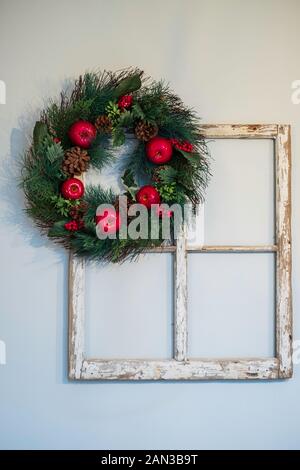 Ein roter apfelweihnachtskranz, der an einem alten Vintage-Fensterrahmen hängt, an der Wand im Innenbereich. Stockfoto