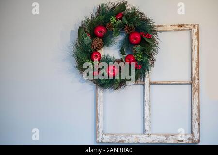 Ein roter apfelweihnachtskranz, der an einem alten Vintage-Fensterrahmen hängt, an der Wand im Innenbereich. Stockfoto