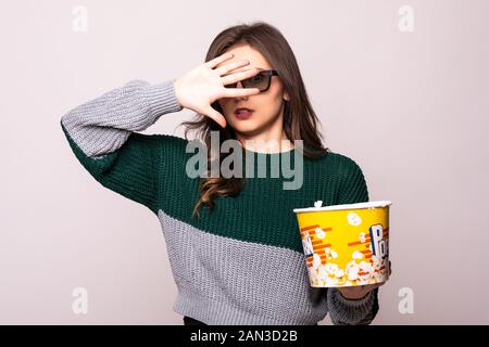 Erschrocktes junges Mädchen in 3D-imax-Brille, die Gesicht mit Handfläche bedeckt, Filmaufnahmen ansieht und einen Eimer Popcorn auf weißem Hintergrund hält. Stockfoto