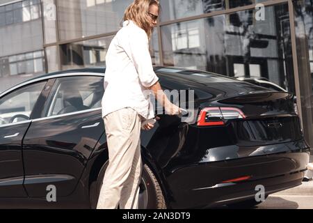 Transport. Junger Mann mit langen Haaren in der Sonnenbrille von Electric Auto stop an chraging Station einfügen plug Reisen Stockfoto