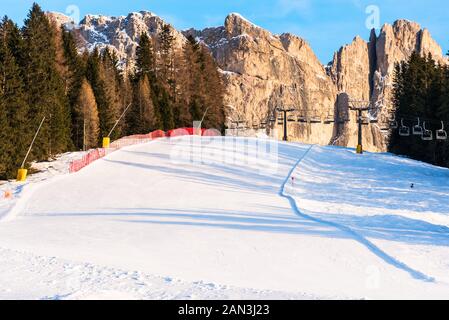 Einsame Piste mit einem Sessellift im Hintergrund am Fuße der hoch aufragenden felsigen Gipfeln der Dolomiten auf einem klaren Winter am Nachmittag Stockfoto