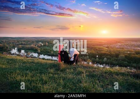 Man Touristen mit Rucksack sitzt auf Gras und Blick vom Berg Hügel zu Sonnenuntergang, der Tourist Rest auf einem Berg, die Freude an der Natur und den Sonnenuntergang. Adven Stockfoto