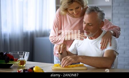 Schöner Mann schneidet Gemüse, fürsorgliche Frau ihn umarmen, Ehepartner Unterstützung Stockfoto