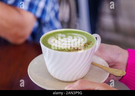 Hand der Person nehmen Sie eine Tasse heißen grünen Teelatte mit Blumenmuster aus Latte Art in weißer Tasse. Hiphster vibes und Vintage-Ton. Stockfoto