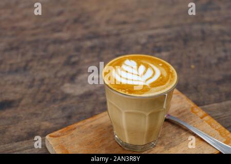 Eine Tasse Cappuccino oder Latte mit Blumenmuster aus Latte Art in transparentem Glas auf Holztablett und Tisch. Hiphster vibes und Vintage-Ton. Stockfoto