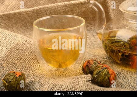 Blumen Tee in eine Teekanne aus Glas gebraut, ein Glas Blume Kaffee und Kugeln von flower Tee auf dem Hintergrund der rauen handgewebte Gewebe. Nahaufnahme Stockfoto