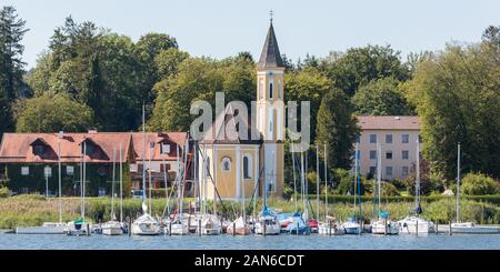 Blick auf die Kirche St. Alban und den Jachthafen mit Segelbooten am Ammersee. Bäume im Hintergrund. Schöne, idyllische Landschaft oberbayerns. Stockfoto