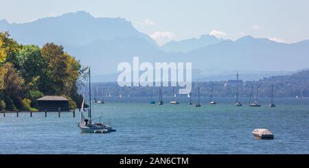 Panoramasicht auf den Ammersee - mit Segelbooten und einem Bootshaus im Vordergrund. Im hinteren Marienmuenster Diessen und die bayerischen alpen. Stockfoto