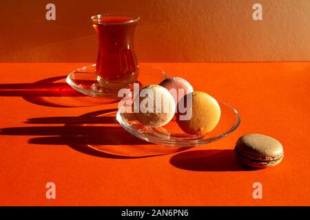 Traditionelle türkische Glas Kaffee mit Süßigkeiten wirft einen dunklen Schatten. Französische desserts Macarons. Minimale Zusammensetzung auf rot-orange Farbe Hintergrund. Stockfoto