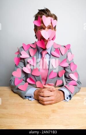 Büroangestellter in Rosa flatternden Herzen sitzend mit gefalteten Händen bedeckt an seinem Schreibtisch Stockfoto