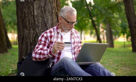 Älterer Mann sitzt auf Gras und bei der Buchung von Tickets online, männlich Ruhe im Park Stockfoto