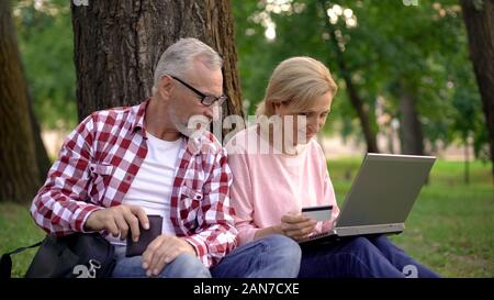 Glückliches Paar sitzt auf Gras und der Auswahl neuer Güter auf Laptop, online einkaufen Stockfoto