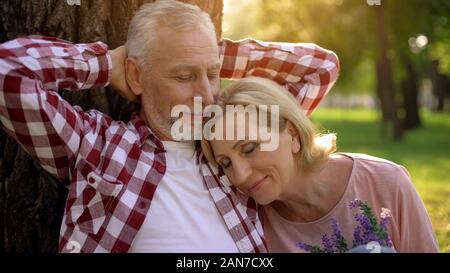 Altes Ehepaar sitzt im Park in der Nähe von Tree, liegende Frau auf mans Schulter, dating Stockfoto