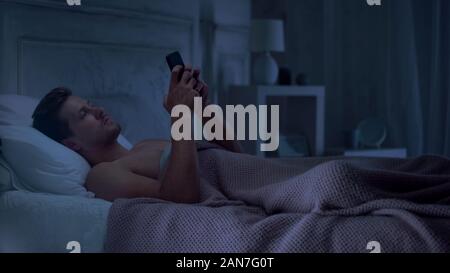 Sleepy männlichen liegend auf Bett und über Handy, Einstellung Alarm für Morgen Stockfoto