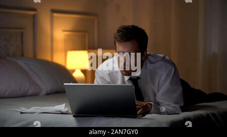 Nachdenklich männlichen liegend auf Bett und Lesen von Dokumenten auf Laptop, e-mail Stockfoto