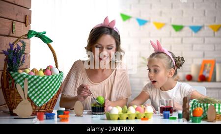 Kleines Mädchen aufgeregt beobachten Mutter eintauchen Ei in grüne Lebensmittelfarbe, Ostern Stockfoto