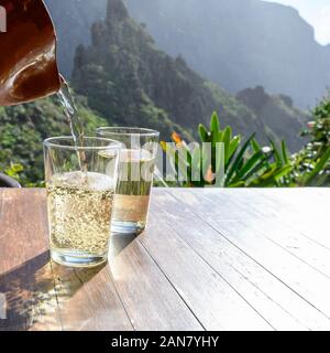 Man gießt Weißwein aus Ton Kanne in Glas auf der Terrasse mit Blick auf die grüne Landschaft der kleinen Bergdorf Masca auf Teneriffa, Spanien im sonnigen d Stockfoto