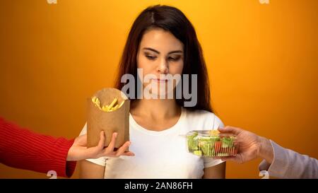 Junge Frau die Wahl zwischen frischem Salat und Pommes frites, zögern zu entscheiden, Stockfoto