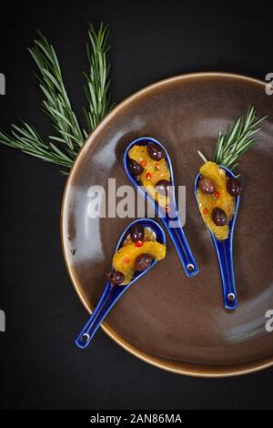 Vegan gesundes Essen. Canape von Orangen, Oliven, Chili und Rosmarin, serviert auf einem keramischen Löffel mit einem schwarzen Hintergrund. Stockfoto