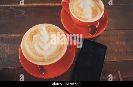 Zwei rote Tassen Kaffee mit latte Kunst auf Holz- Desktop mit dem Smartphone. Tabelle im Cafe. Ansicht von oben. Stockfoto