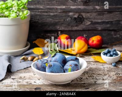 Herbst Hintergrund mit Reifen violett blau Pflaumen in Weiß Schüssel auf alten grauen Holztisch, Seitenansicht Stockfoto