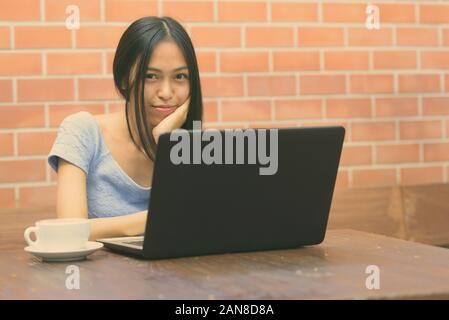 Junge schöne asiatische Jugendmädchen ruhenden Kinn auf Hand mit Laptop und Cappuccino auf hölzernen Tisch gegen die Wand Stockfoto