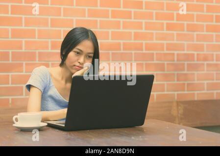 Junge schöne asiatische Jugendmädchen ruhenden Kinn auf Hand, während Sie mit Laptop mit Cappuccino auf hölzernen Tisch gegen die Wand Stockfoto