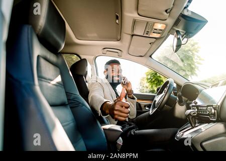 Erfolgreiche schöner afrikanischer Mann Geschäftsmann und Chef im Auto sitzen, telefonieren, Lächeln, schauen in die Kamera und zeigen Daumen hoch Stockfoto