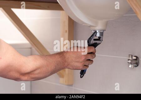 Die Hand des Closeup männlichen Klempner zur Festsetzung Waschbecken im Bad mit Fliesen an der Wand. Professionelle sanitär Reparatur, Installation von Wasserleitungen. In den Händen Schraubenschlüssel Stockfoto