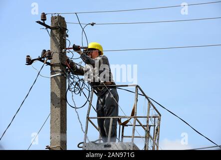 Electrician repairman is working on aerial work platform near top of concrete utility pole with many electric wires of exterior power line on blue sky Stockfoto