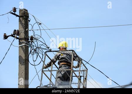 Electrician repairman is working on aerial work platform near top of concrete utility pole with many electric wires of exterior power line on blue sky Stockfoto