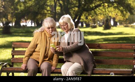 Alte Dame Übersicht News Feed auf Handy zu Freund, sitzt auf der Bank im Park Stockfoto