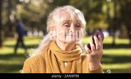 Reife Frau auf ihr Spiegelbild in kompakten Spiegel im Park sitzen auf der Suche Stockfoto