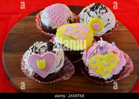 Liebe Muffins, bunten Cupcakes mit Marzipan Herzen, auf Holz- runde Untertasse über den roten Tischdecke serviert dekoriert, Seitenansicht Stockfoto