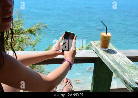 Junge Jugendmädchen Erfrischung im Café mit Meerblick ang Fotografieren ihr Eis Kaffee frappe per Handy Stockfoto