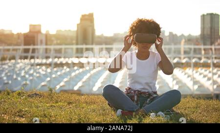 Lustige lockig behaarten Frauen mit VR-Headset, auf Rasen an sonnigen Tag sitzen, Gadget Stockfoto