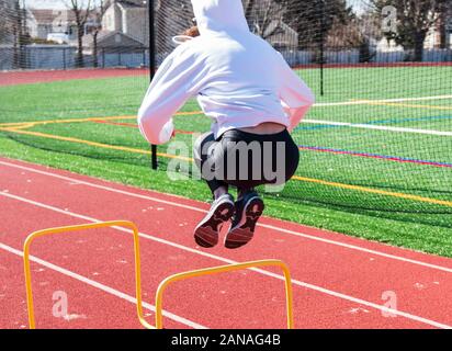 Eine Highschool-Sportlerin mit weißem Hoody und schwarzem Spandex springt während des Geschwindigkeits- und Agilitätstrainings über gelbe Mini-Hürden auf eine Strecke Stockfoto