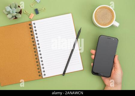 Notebook mit leeren Seite, Bleistift, die sich öffnen lassen, Wasserkocher, Kaffee Tasse und Hand mit einem Smartphone. Table Top, Arbeitsbereich auf grünem Hintergrund. Kreative flach. Stockfoto