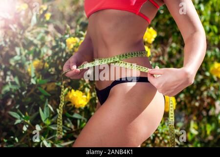 Frau in Rot und Schwarz bikini messen ihre Taille neben grünen Hintergrund. Fitness, Diät und Gesundheit Konzept Stockfoto
