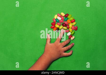 Eine kleine Kinder Hand erreichen für ein Bündel von Bean geformten Süßigkeiten verschiedener Aromen, mit einem grünen Hintergrund. Stockfoto