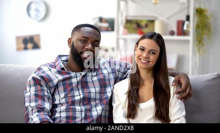 Lächelnd mixed-Race Paar sitzt auf einem Sofa zu Hause und suchen an Kamera, Familie Stockfoto