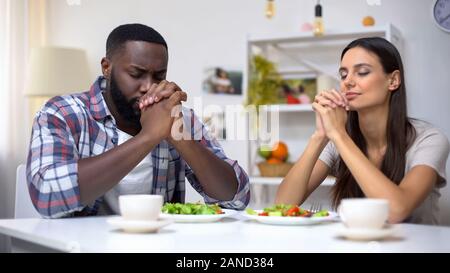 Junge mixed-Race Paar Beten vor dem Essen, vegetarische gesunde Salate auf Tisch Stockfoto